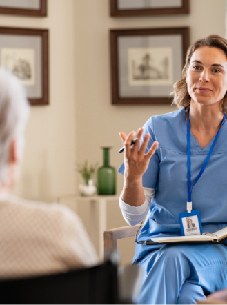 A woman in blue scrubs, holding a clipboard, talks to a patient